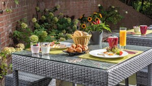 Una mesa de desayuno al aire libre con una mesa de mimbre, platos de comida, una cesta de pan, una planta en maceta y un vaso de jugo de naranja.