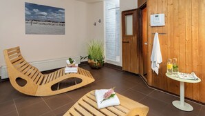 Una habitación con suelo de madera, un sillón mecedor de madera, una mesita con una toalla y botellas, una planta en maceta y un ventanal con vista al cielo.