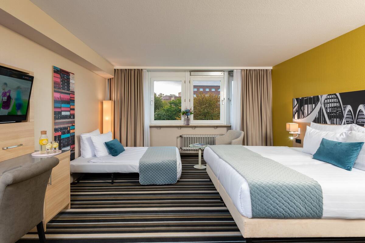Ein Hotelzimmer mit zwei Betten, einem Fernseher, einem Schreibtisch und einem Fenster mit Blick auf Gebäude.