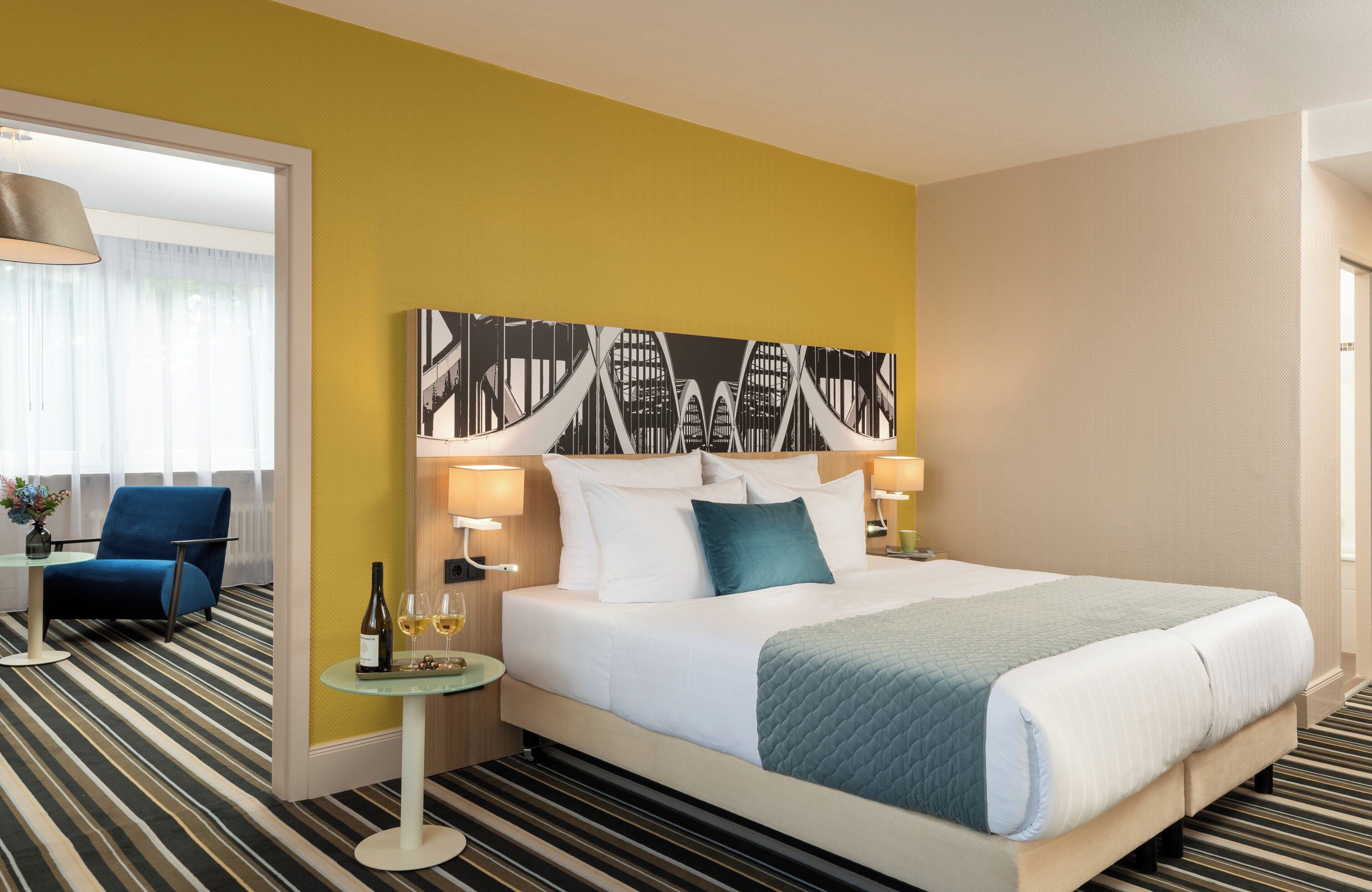 Een hotelkamer met een bed, een blauwe stoel, een klein tafeltje met een fles en glazen, en een schilderij aan de muur.