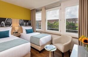 Habitación de hotel con dos camas, una silla, una mesa y un ventanal con vistas a edificios y zonas verdes.