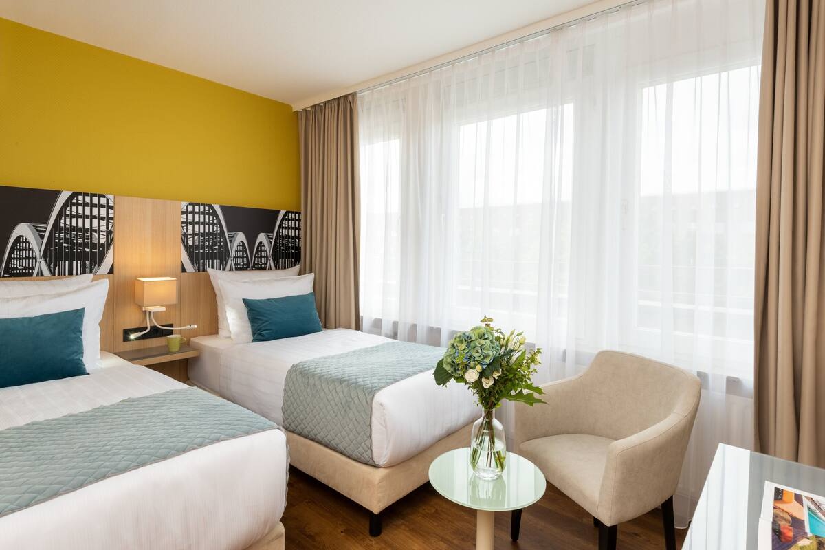 Ein Hotelzimmer mit zwei Betten, einem Sessel, einem kleinen Tisch und einer Vase mit Blumen.