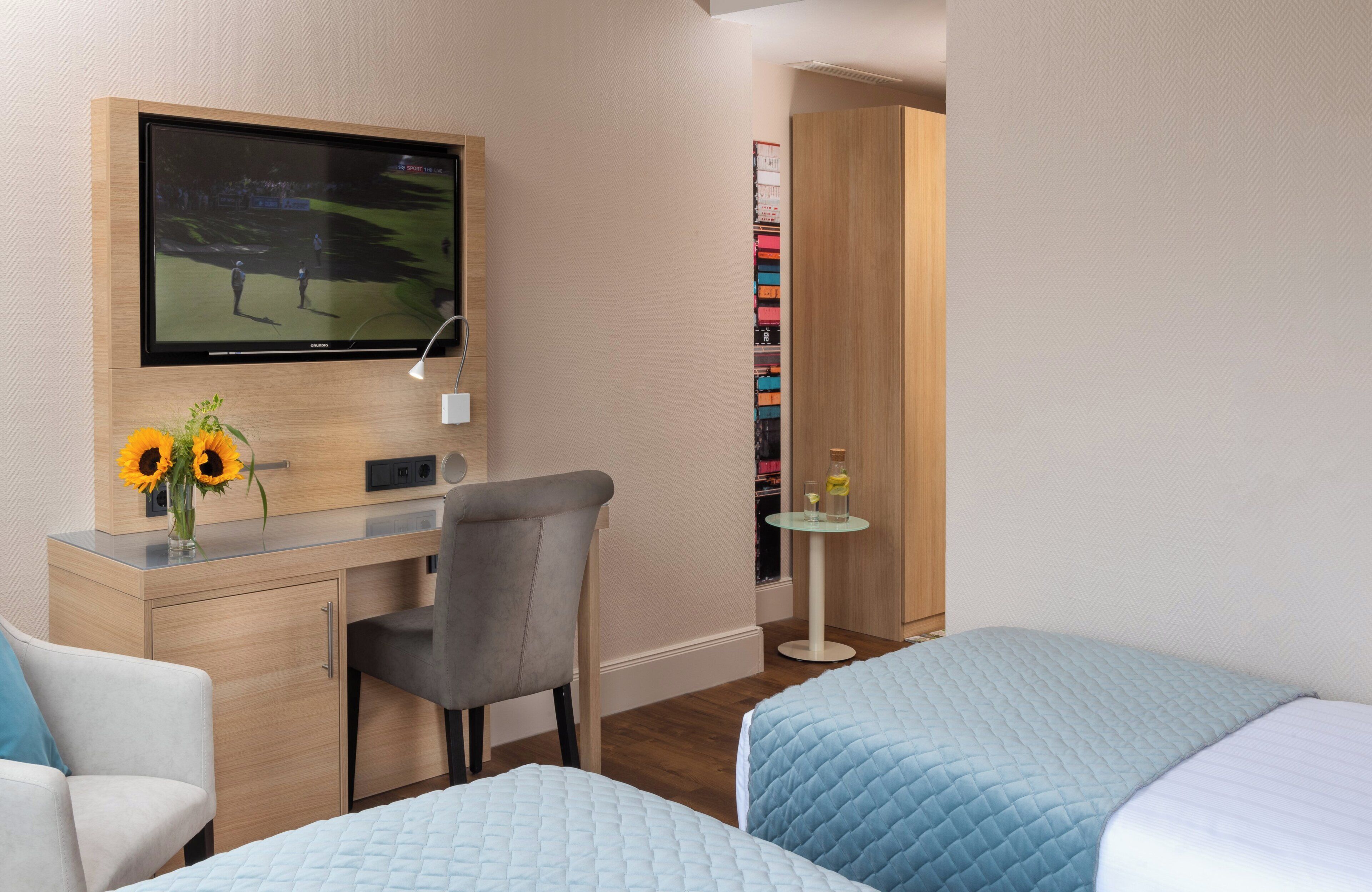 Habitación de hotel con televisor, escritorio con lámpara, silla, dos camas, mesita con botella y vaso, y pared con diseño en espiga.