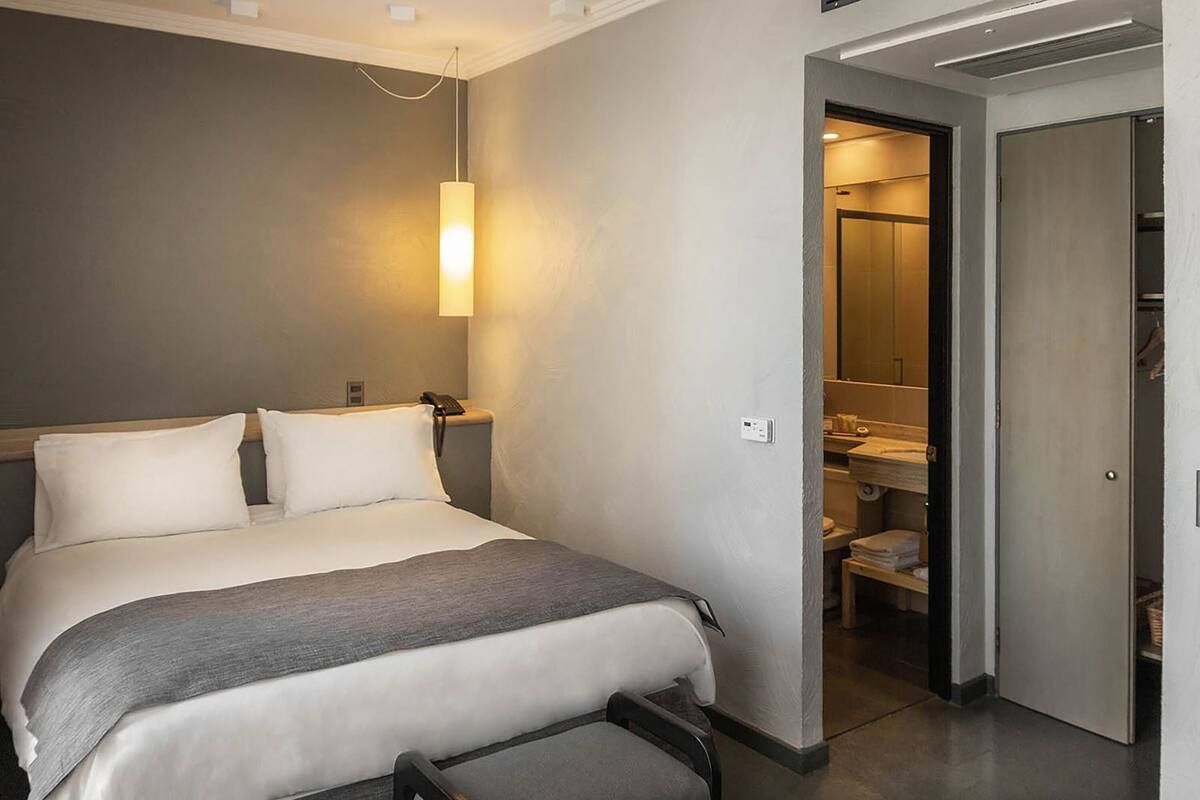 A hotel room with a bed, bedside table, lamp, and a bathroom visible through an open door.