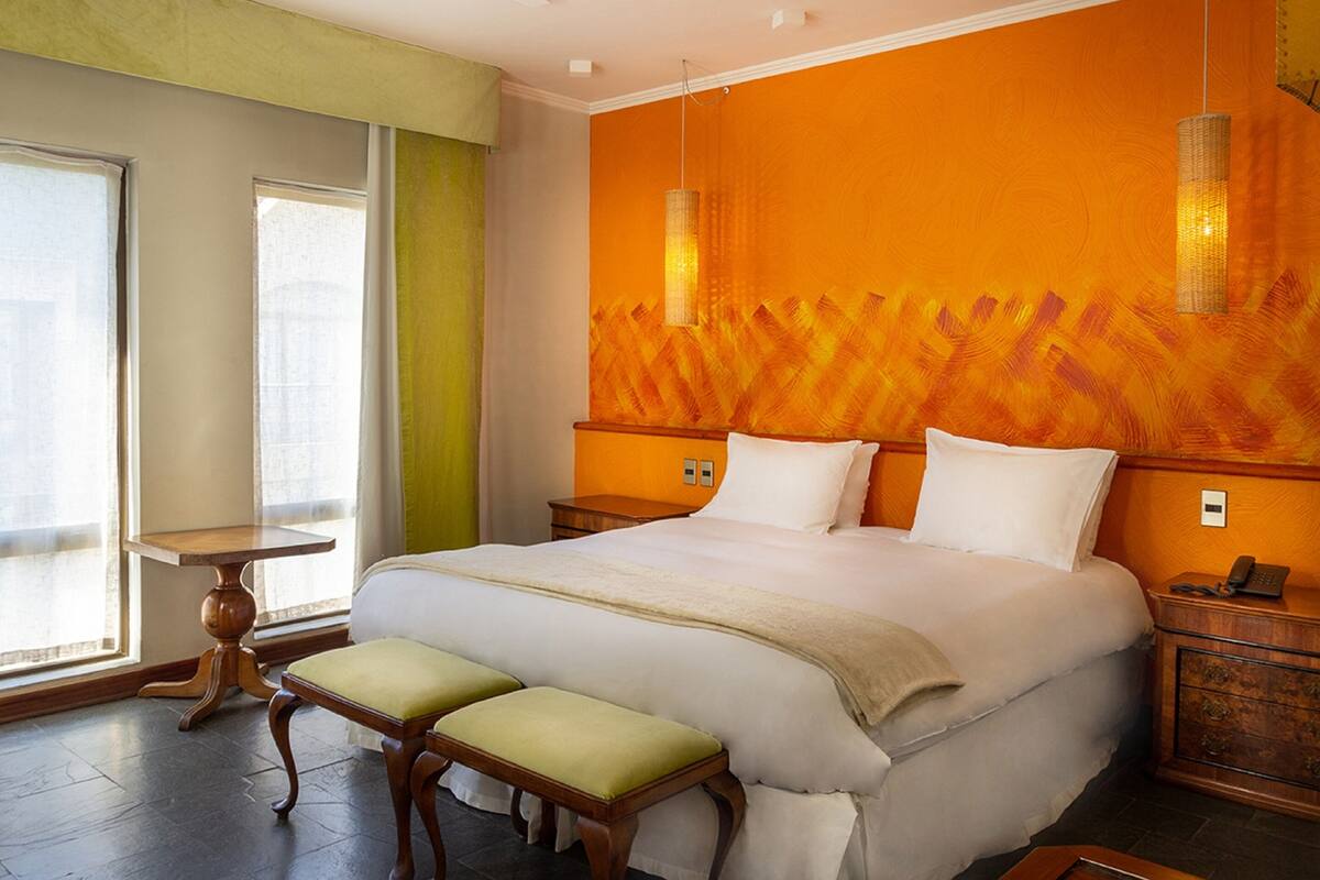 A hotel room with a large bed, a bench, a bedside table, a nightstand, and a window with curtains.