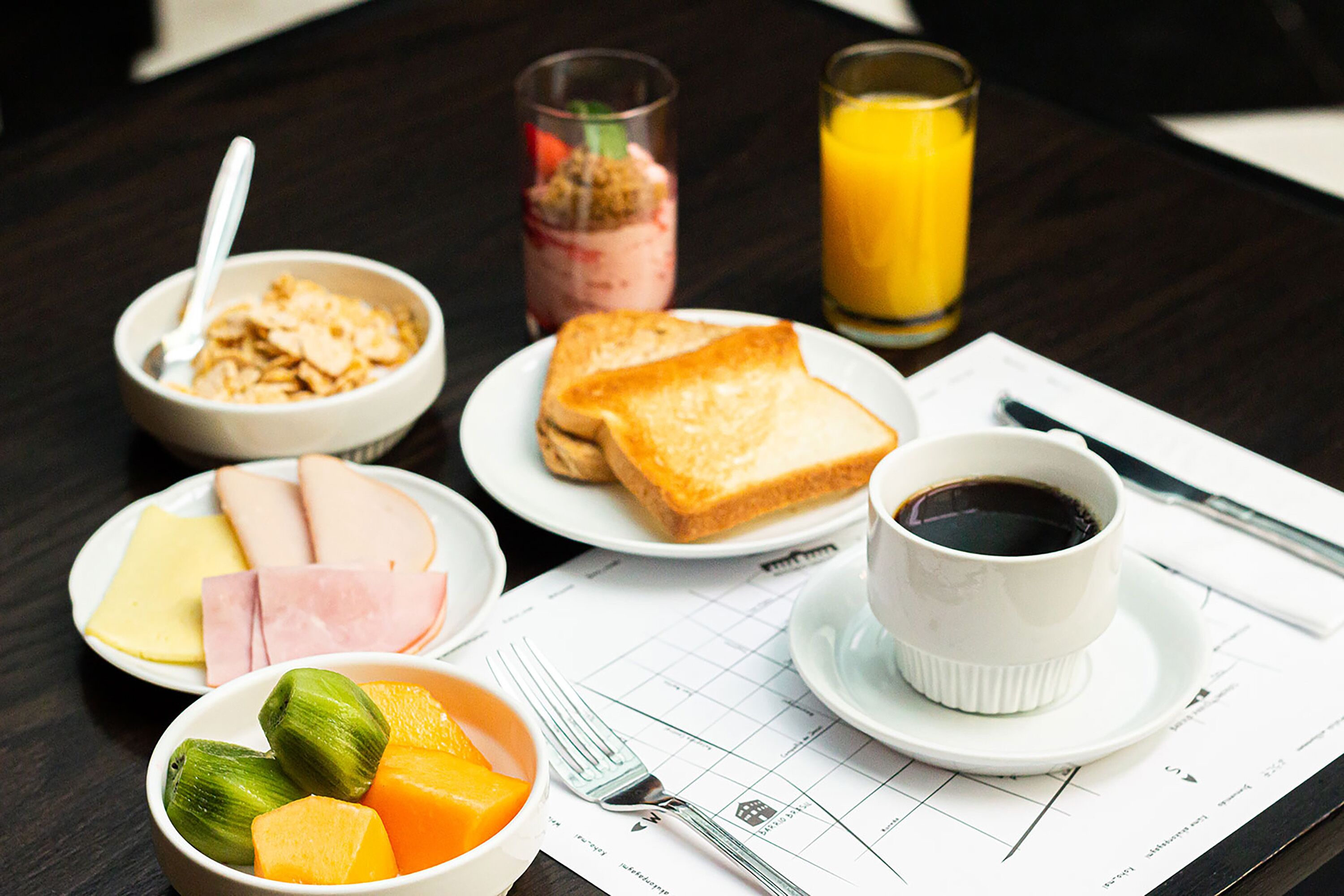 A breakfast table with cereal, toast, ham, cheese, fruit, a cup of coffee, and a glass of orange juice.