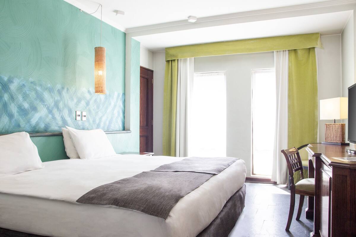 A hotel room with a large bed, a desk with a lamp, and a view of the outdoors through a window with green curtains.