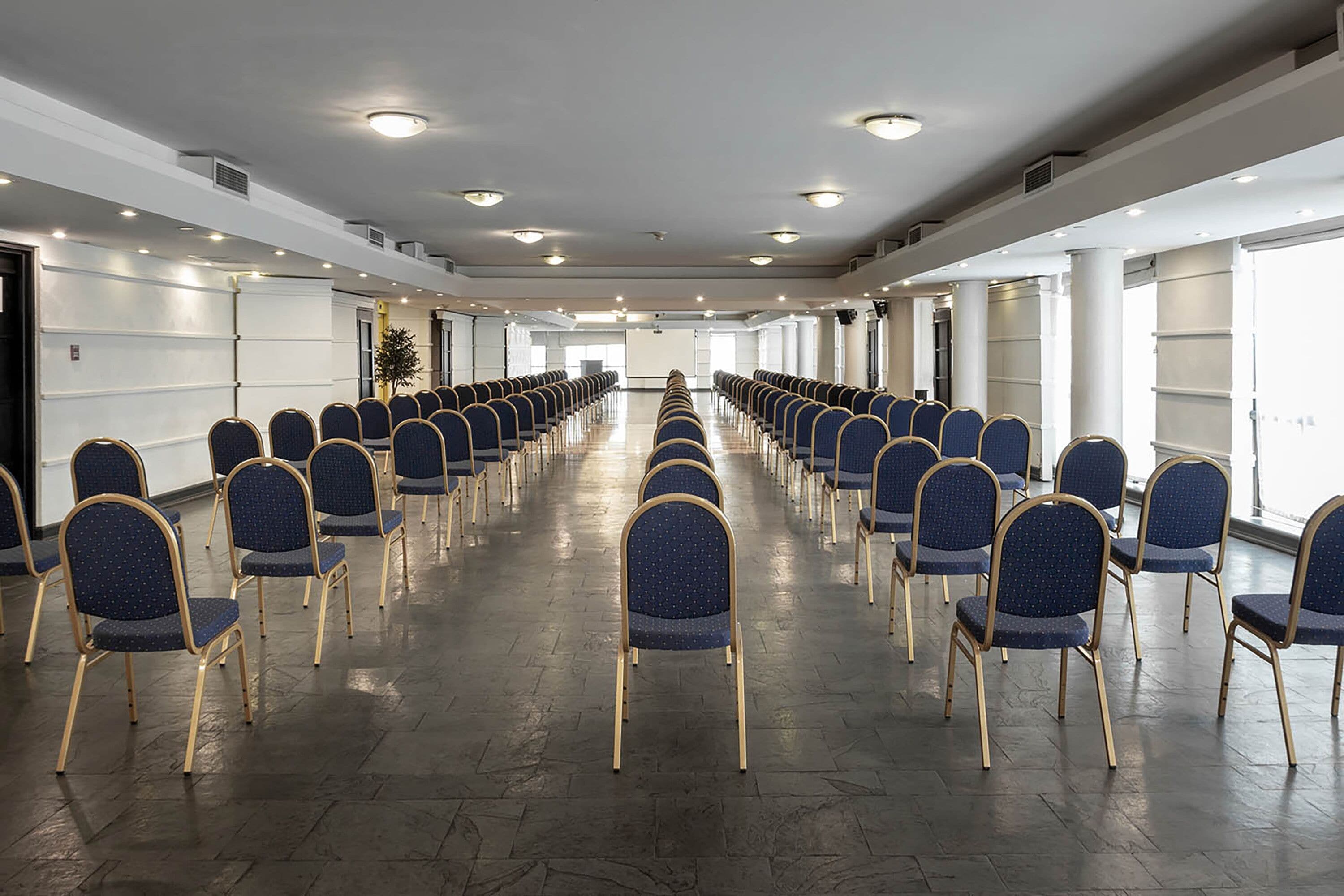 Un salón de conferencias con filas de sillas, ventanas amplias y un escenario al frente.