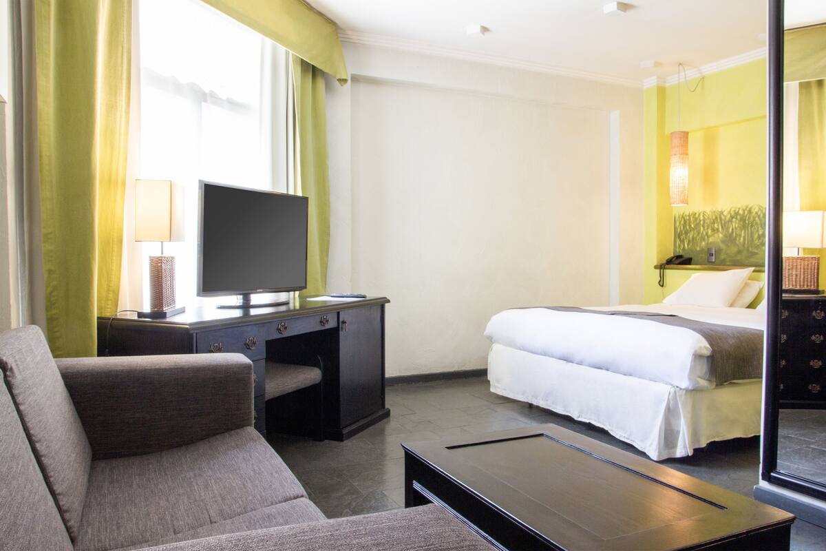 A hotel room with a bed, a desk with a television, a sofa, and a large mirror.