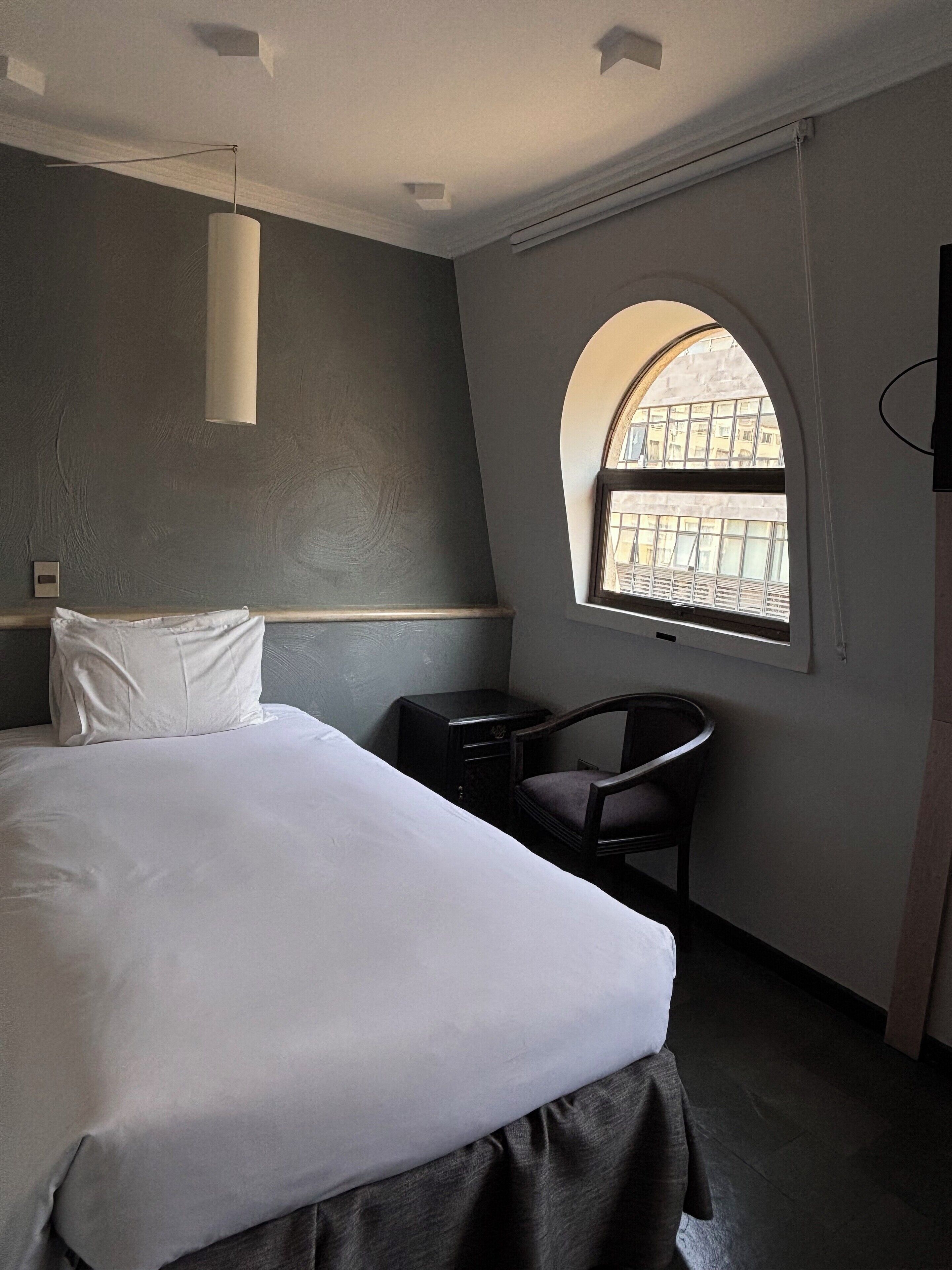 Habitación de hotel con una cama grande, un escritorio pequeño, una silla y un ventanal con vista arqueada.