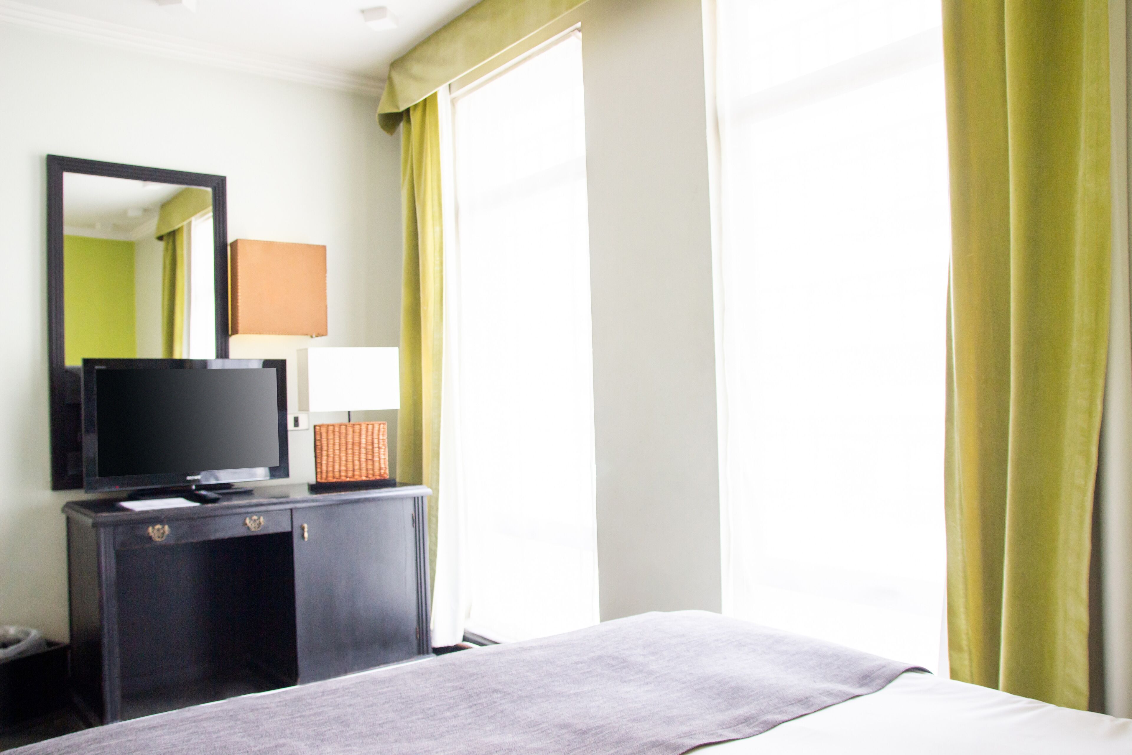 A hotel room with a bed, a TV on a dresser, a window with curtains, and a mirror on the wall.