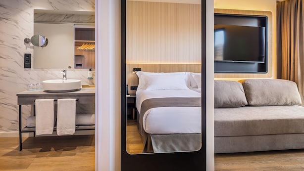 A modern hotel room with a marble bathroom, a bed with grey bedding, and a large TV mounted on the wall.