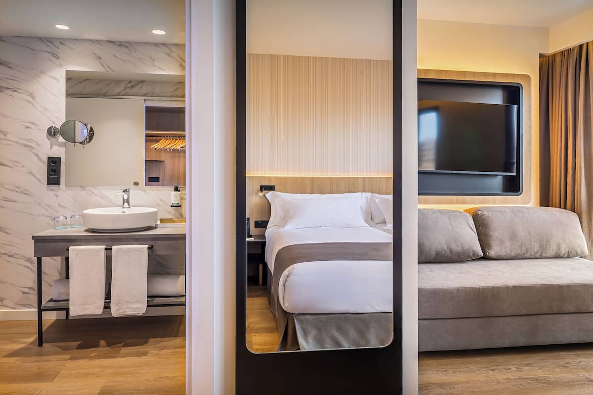 Una habitación de hotel moderna con baño de mármol, cama con ropa de cama gris y un televisor grande montado en la pared.