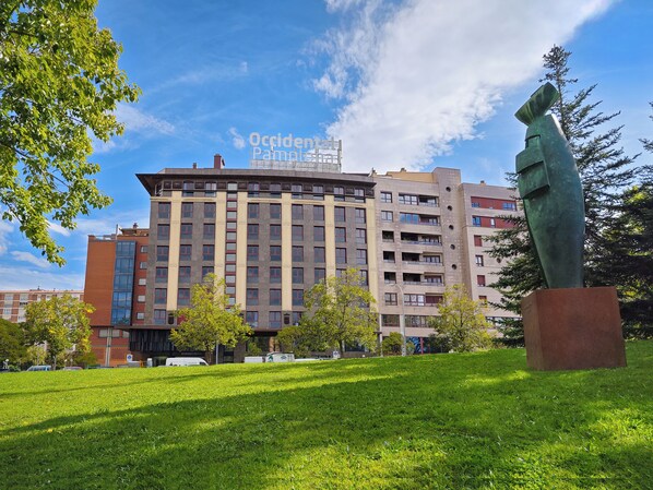 Un edificio de varios pisos con un diseño arquitectónico moderno, que presenta una combinación de ventanas rectangulares y curvas, y una escultura destacada en primer plano.