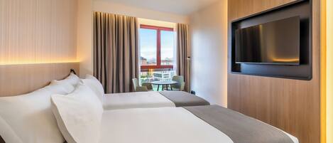 Habitación de hotel con dos camas, un televisor montado en la pared, un balcón con cortinas y vista a edificios.