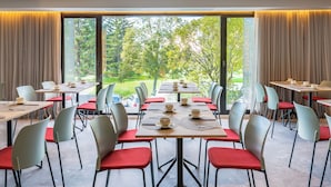 A dining area with multiple tables and chairs, large windows with curtains, and a view of greenery outside.