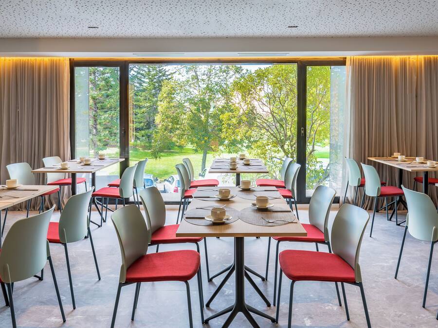 A dining area with multiple tables and chairs, large windows with curtains, and a view of greenery outside.