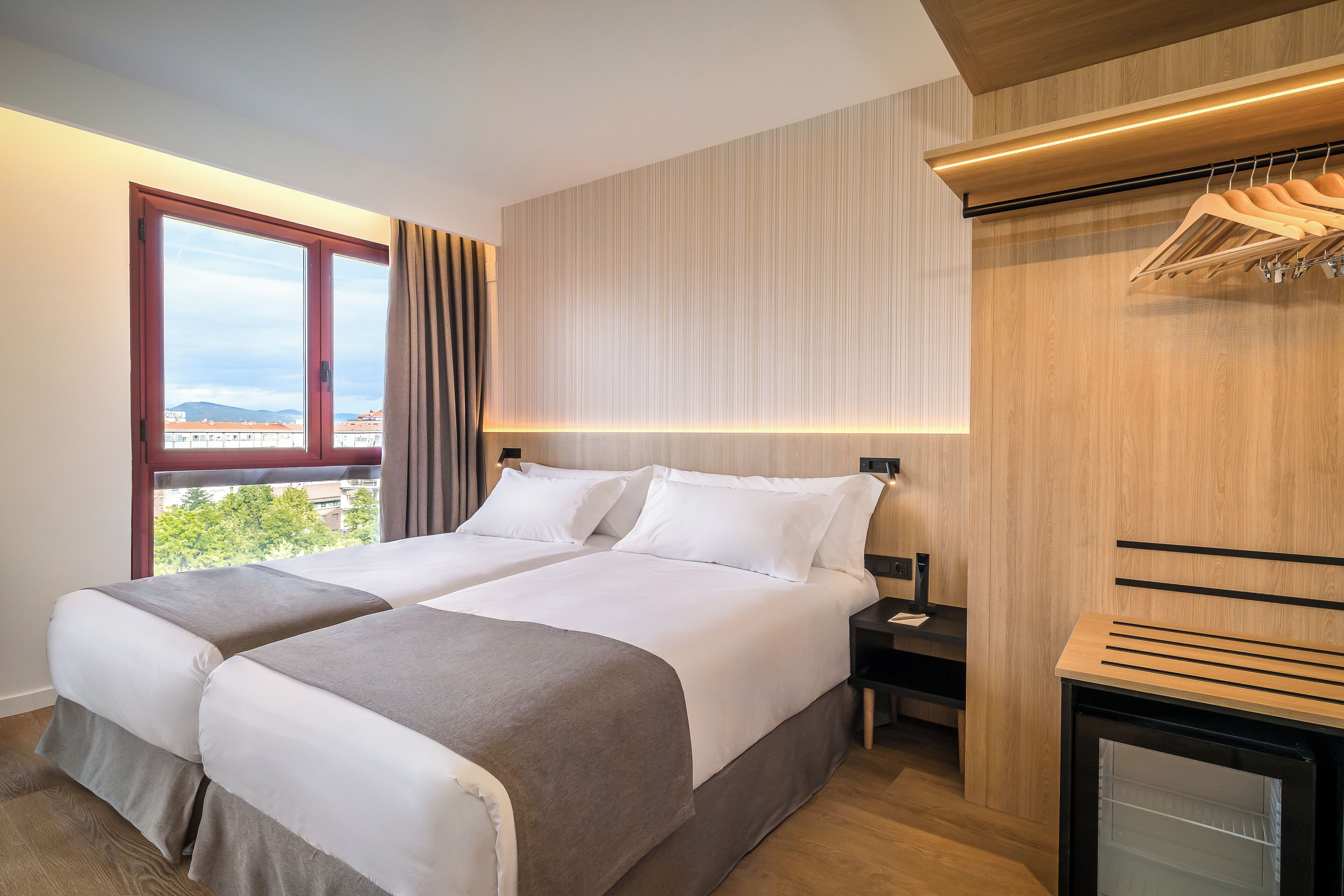 Habitación de hotel con dos camas, un armario de madera, un escritorio pequeño y vistas a edificios a través de una ventana.