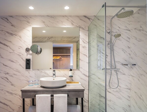 A modern bathroom with a marble wall, a round sink, a glass-enclosed shower, and a mirror with a wooden door in the background. - Occidental Pamplona (Pamplona)
