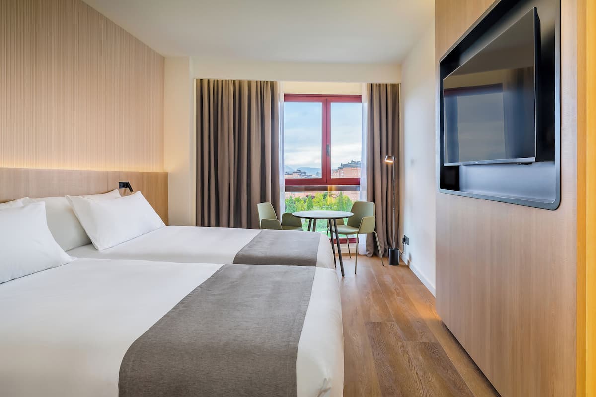 Habitación de hotel con dos camas, televisión, balcón con mesa y sillas, y vista a la ciudad.
