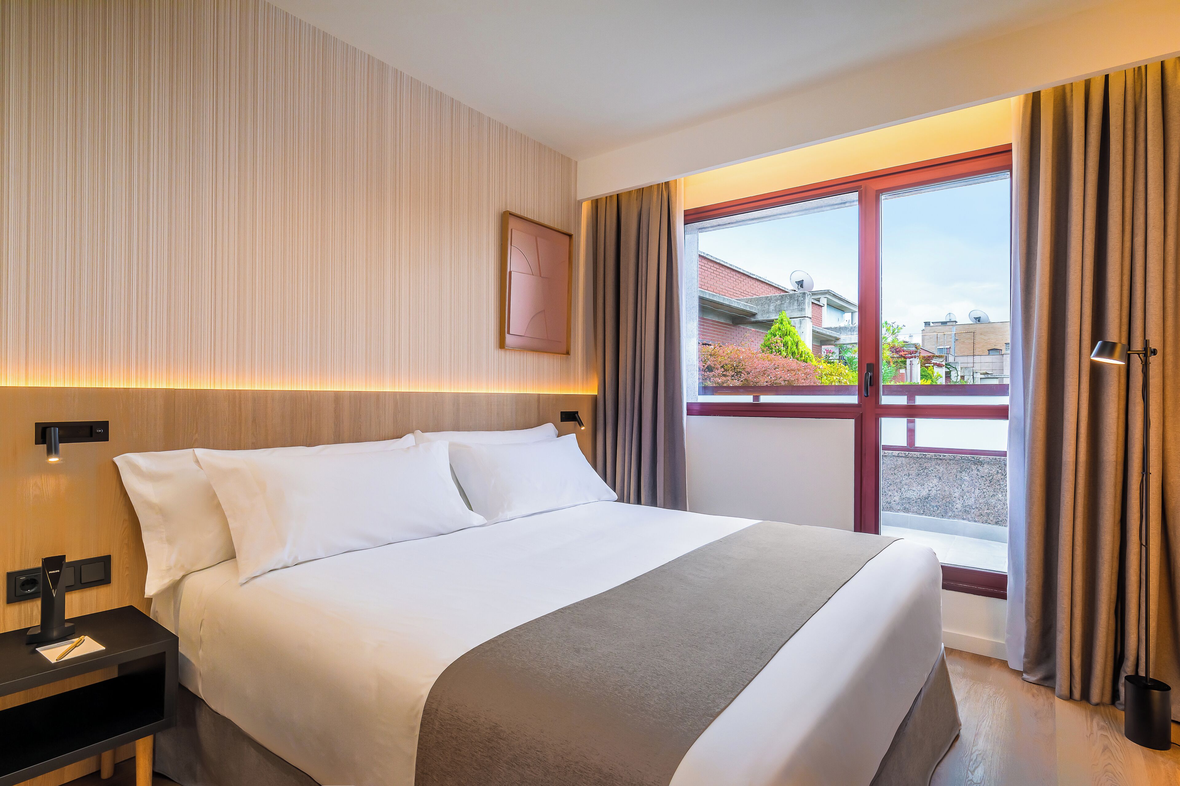 Ein Hotelzimmer mit einem großen Bett, einem Nachttisch, einem Fenster mit Vorhängen und Blick auf Gebäude.