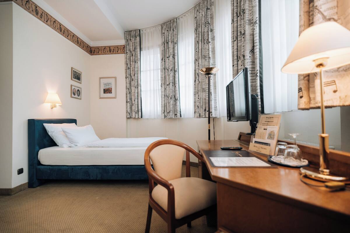 A hotel room with a bed, a desk with a lamp, a chair, a TV, and curtains with a musical note pattern.