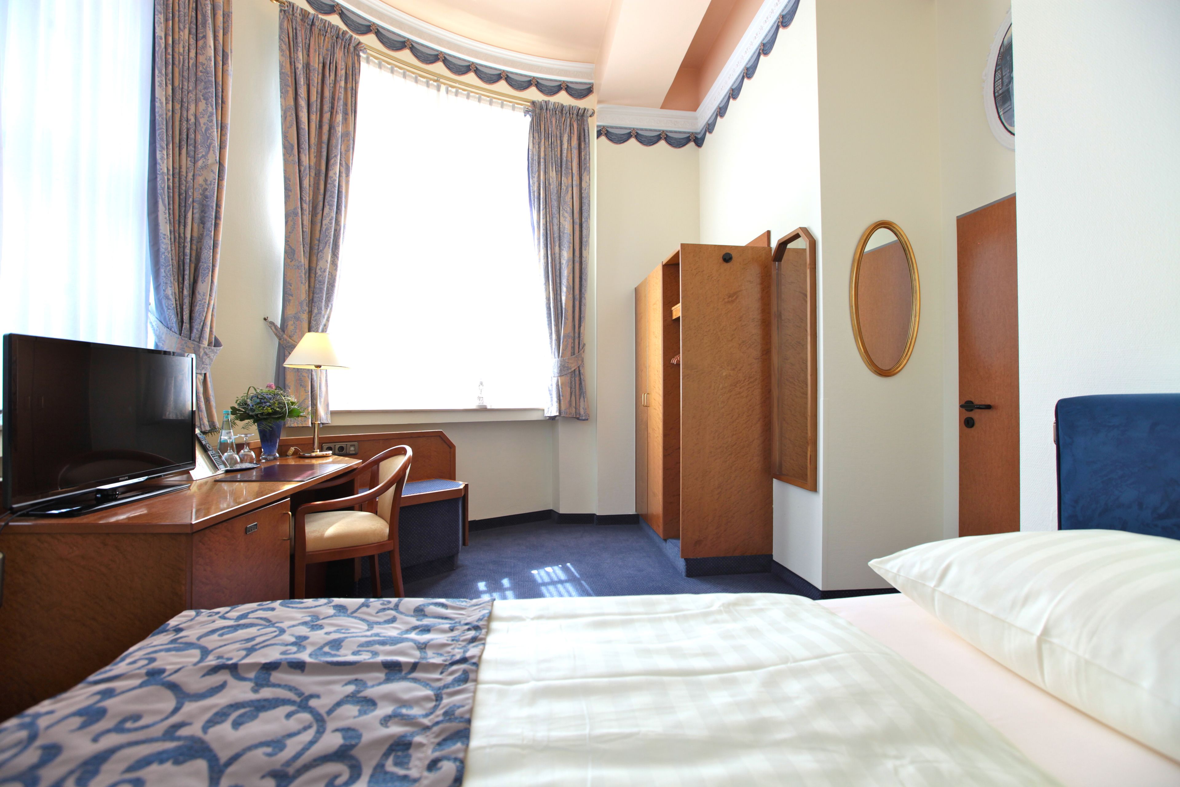 A hotel room with a bed, a desk with a TV, a chair, a desk lamp, and a window with curtains.