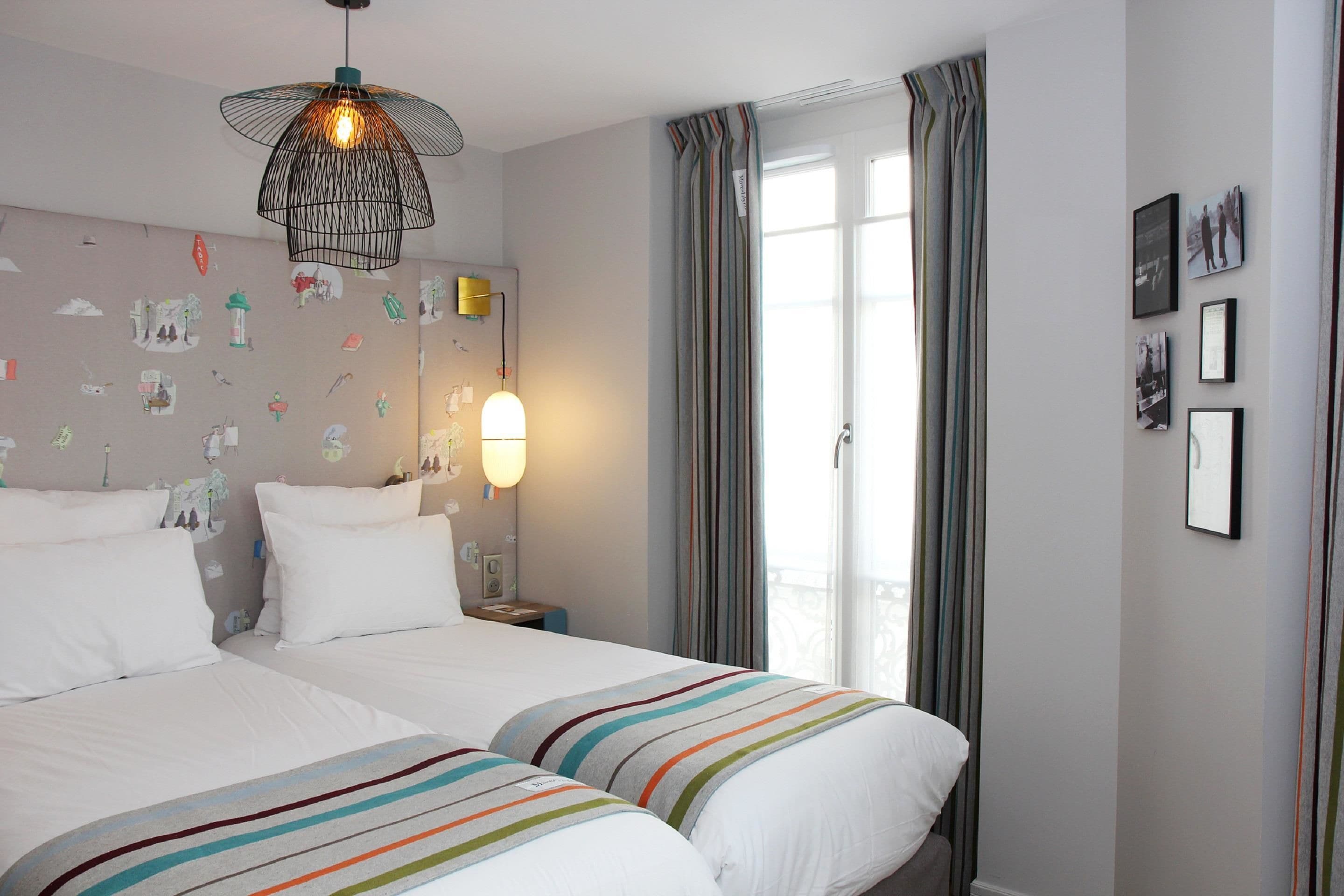 A hotel room with two beds, a wall-mounted lamp, a ceiling light, and a window with curtains.
