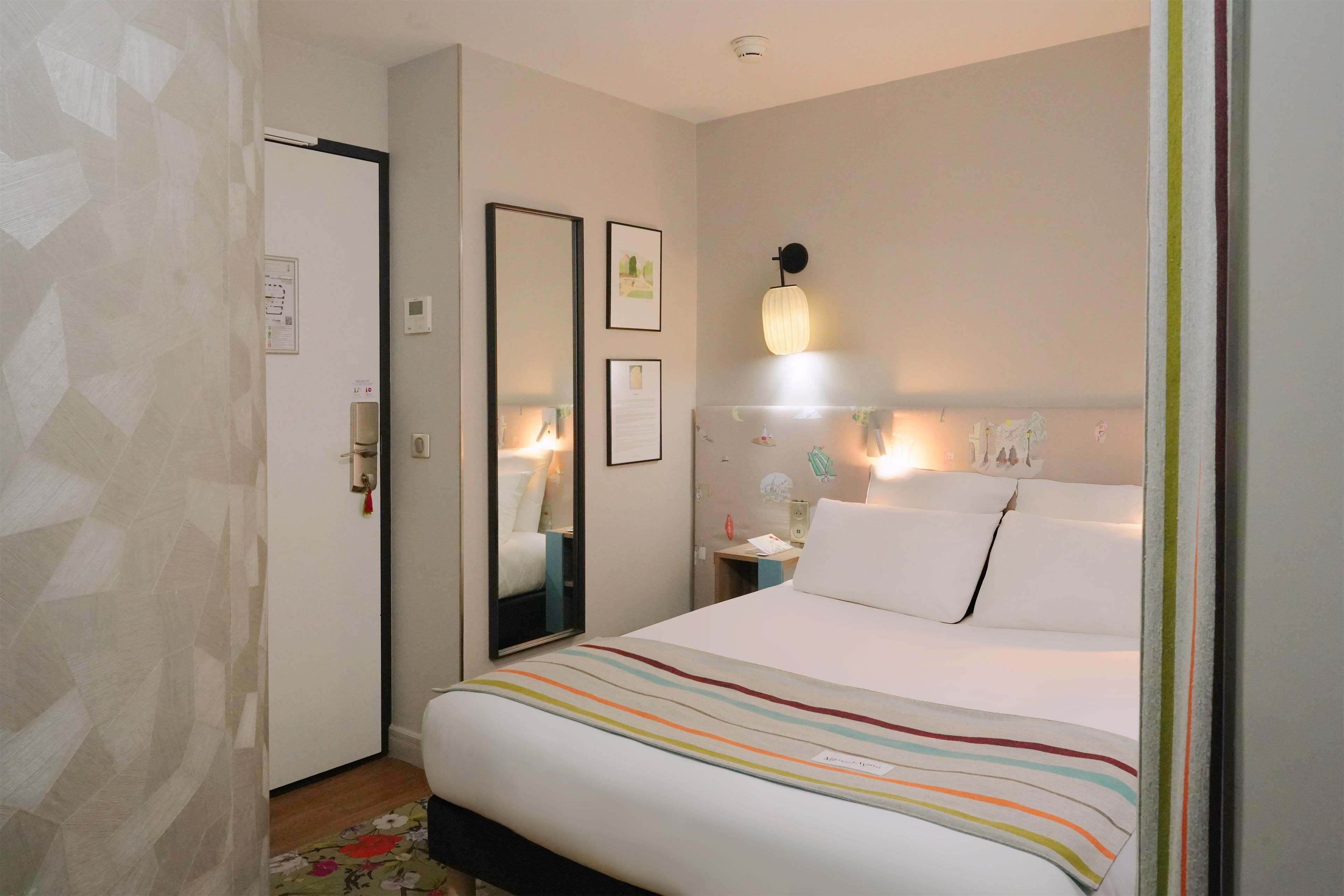 Een hotelkamer met een bed, nachtkastje, spiegel en een lamp aan de muur.