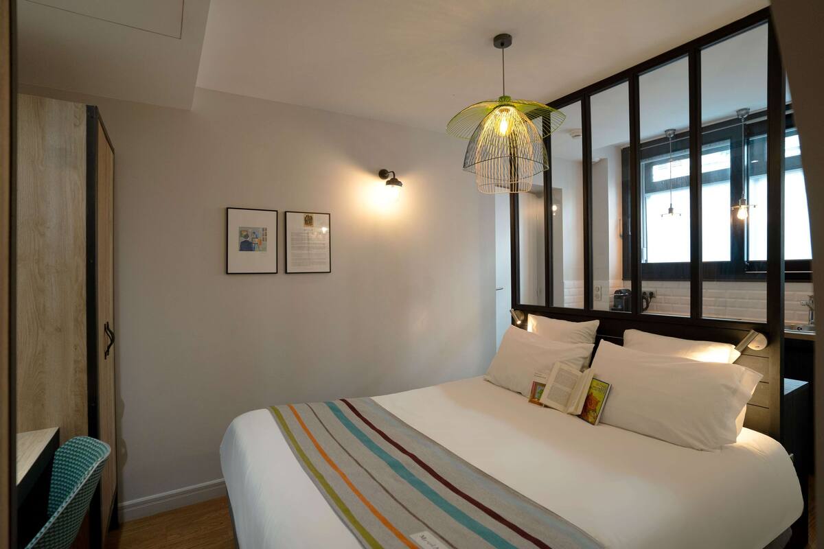 A neatly made bed with a striped blanket, a bedside lamp, and a window with blinds.