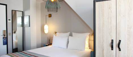 A neatly made bed with white linens, a colorful striped blanket, a modern pendant light, and a wooden wardrobe with black handles.