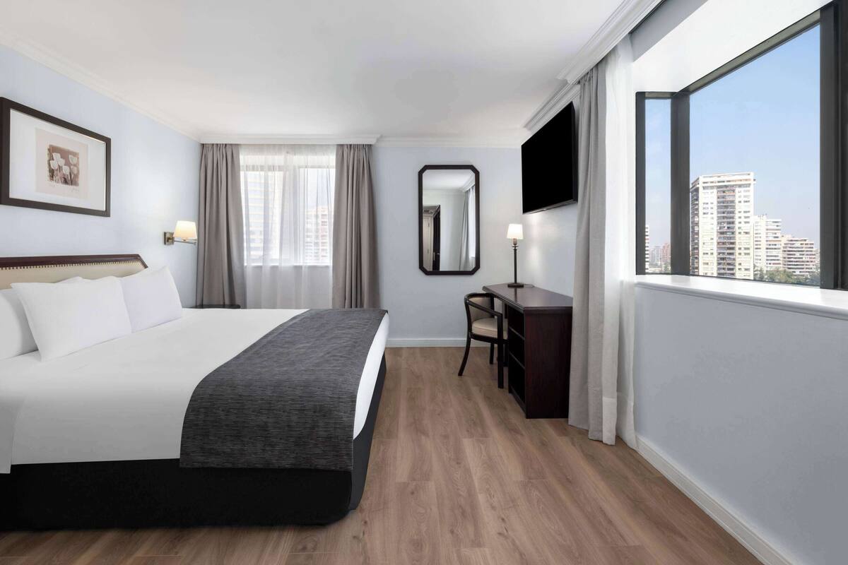 Habitación de hotel con una cama grande, un escritorio, una silla, un televisor de pantalla plana, un ventanal con vistas a la ciudad y un cuadro en la pared.
