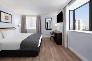 A hotel room with a large bed, a desk, a chair, a flat-screen TV, a window with a city view, and a framed picture on the wall.