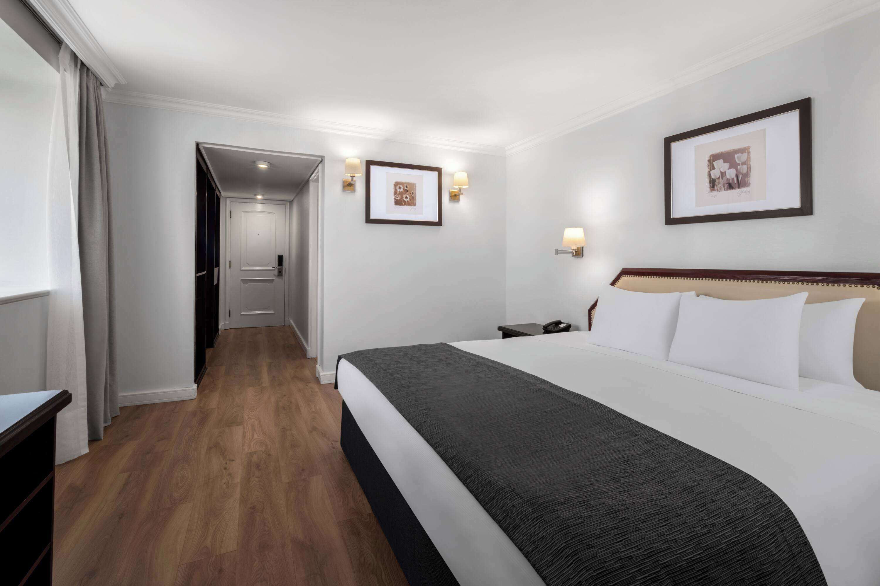 A hotel room with a large bed, a bedside table, a nightstand, a framed picture on the wall, and a door leading to another room.