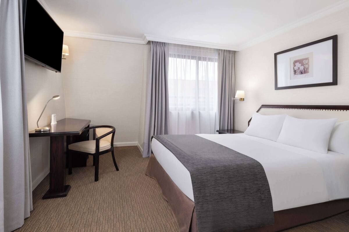 Habitación de hotel con cama, escritorio, silla, televisión y ventana con cortinas.