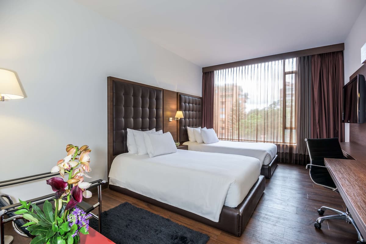 A hotel room with two beds, a desk, a chair, a TV, and a window with curtains.