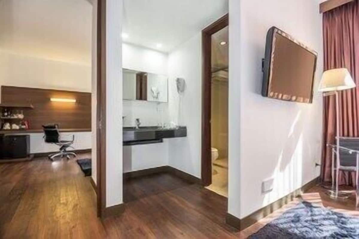A hotel room with a wooden floor, a desk, a chair, a bed, a bathroom with a sink and mirror, and a TV mounted on the wall.