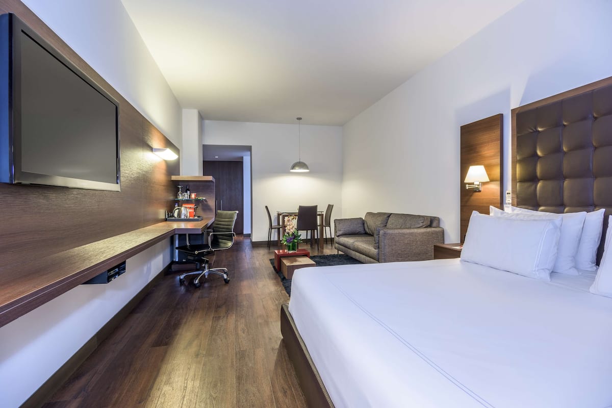 A hotel room with a large bed, a desk with a chair, a sofa, a TV mounted on the wall, and a lamp.
