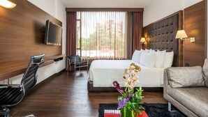 A hotel room with a large bed, a flat-screen TV mounted on the wall, a sofa, a chair, and a small table with a vase of flowers. - NH Collection Bogotá Andino Royal (Bogotá)