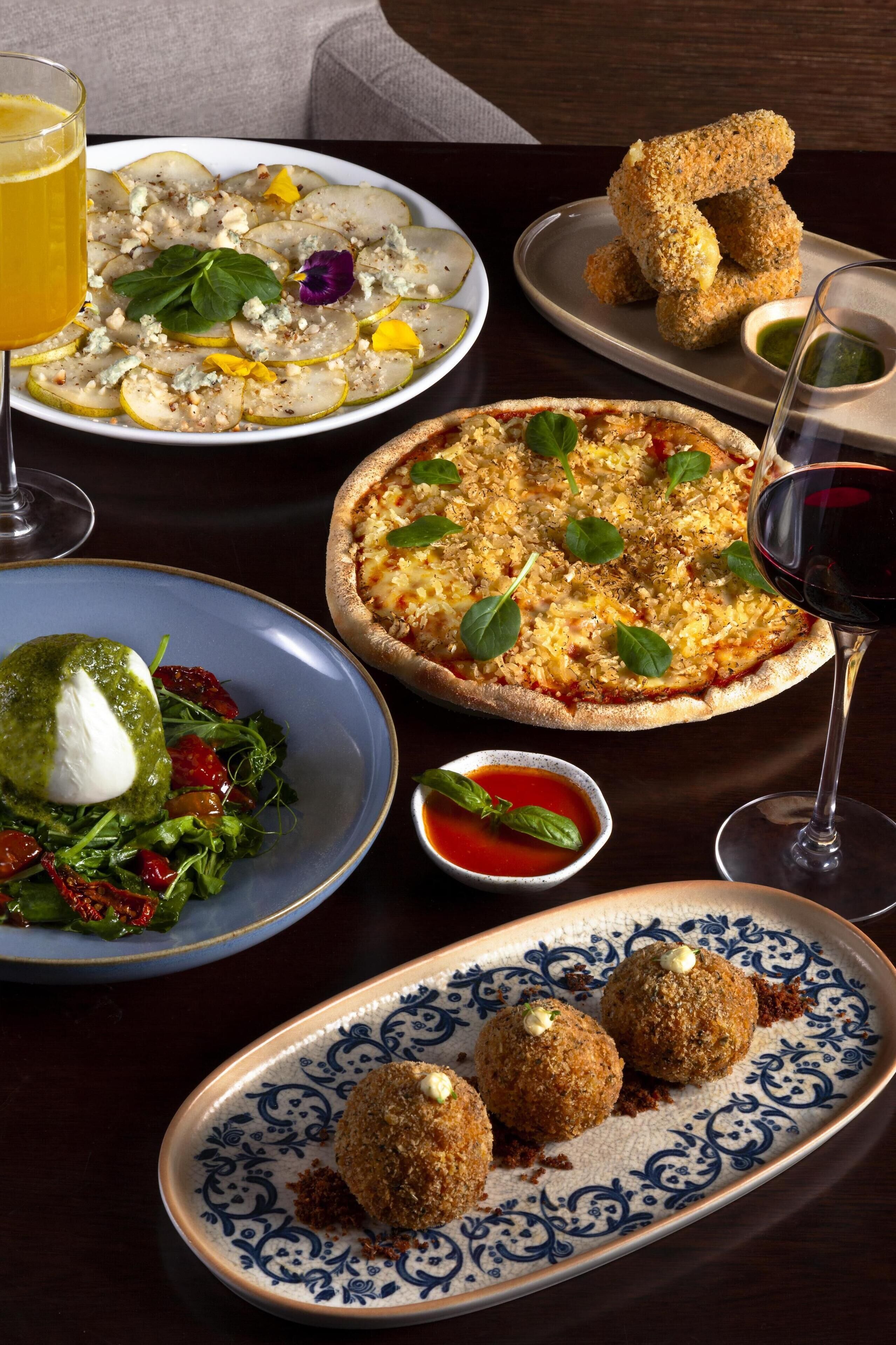 Una variedad de platos que incluyen pizza, croquetas y una ensalada, servidos en una mesa con copas de vino.