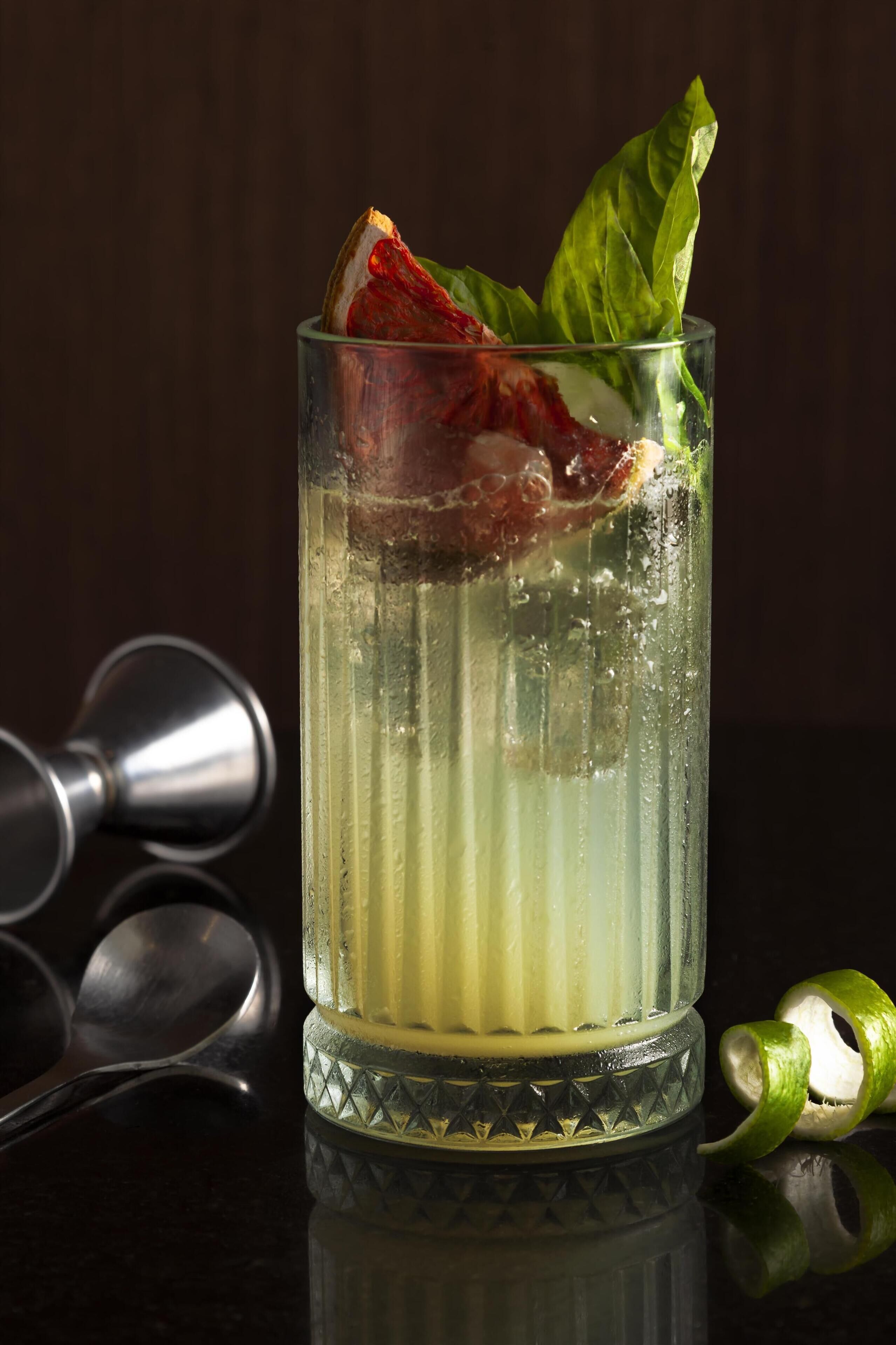 A tall glass with a layered drink, garnished with a leaf and a slice of red fruit, on a reflective surface with spoons nearby.