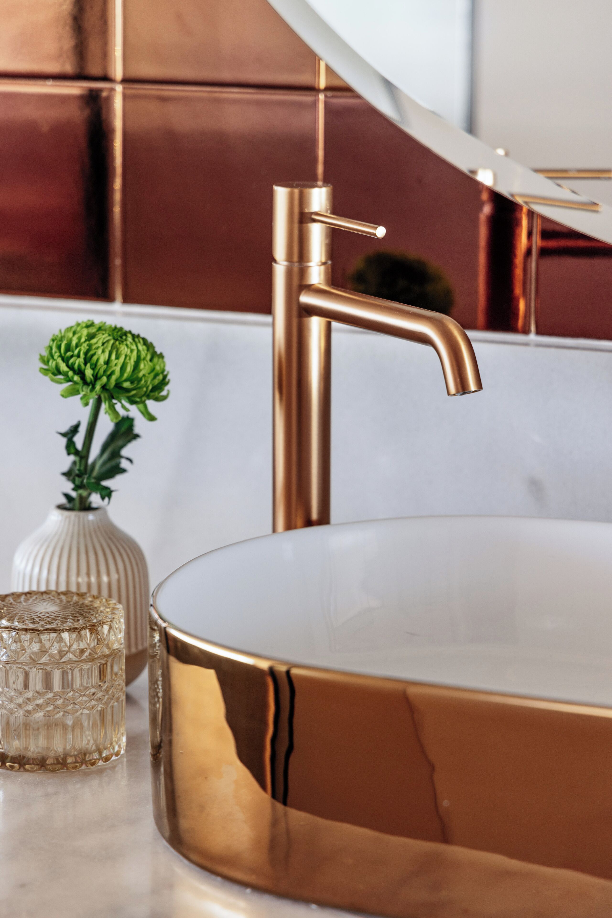 Een moderne badkamer met een gouden badkuip, een glazen vaas met groene bloemen en een decoratieve glazen container.