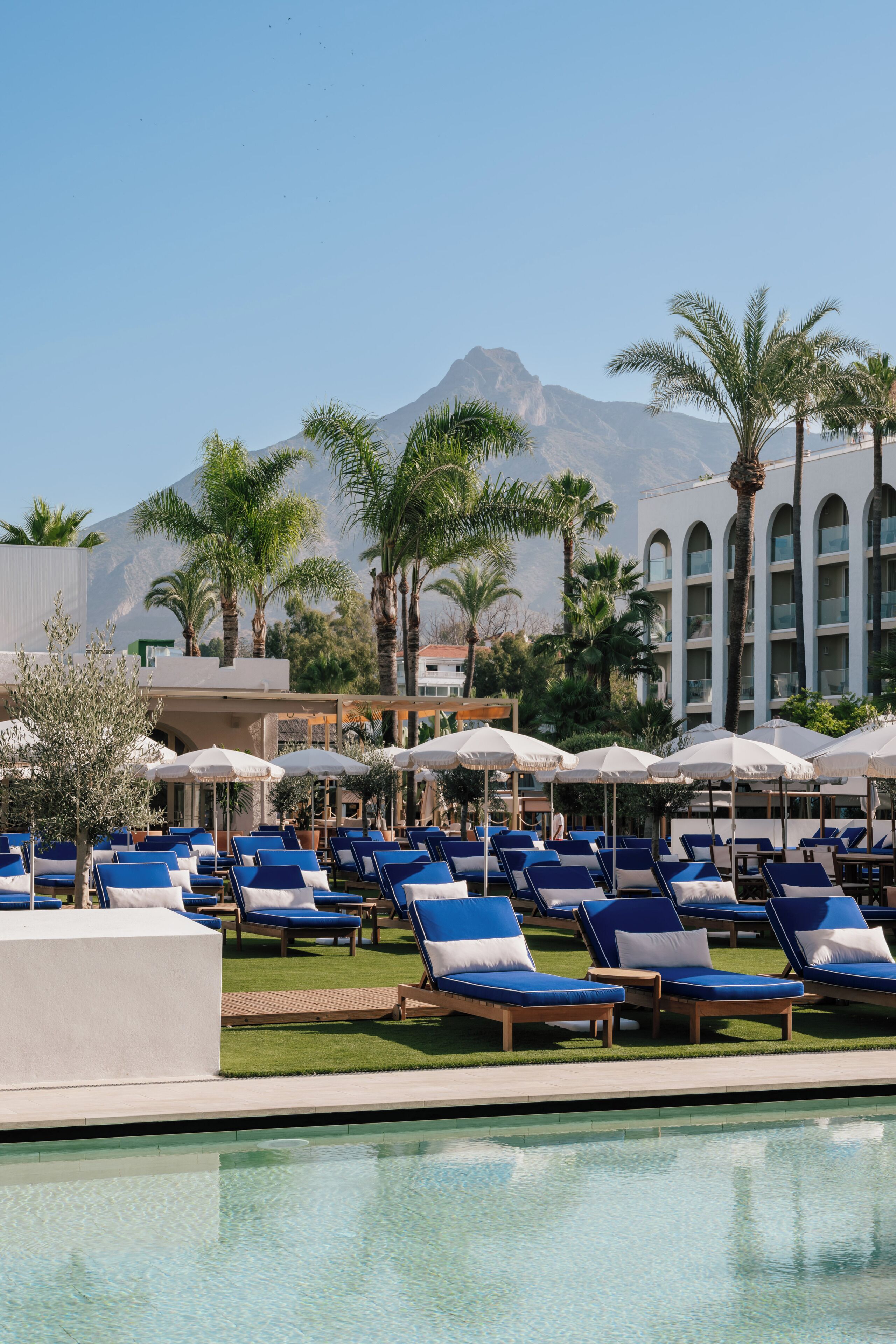 A poolside area with lounge chairs and umbrellas, a building with arched windows, and a mountain in the background.
