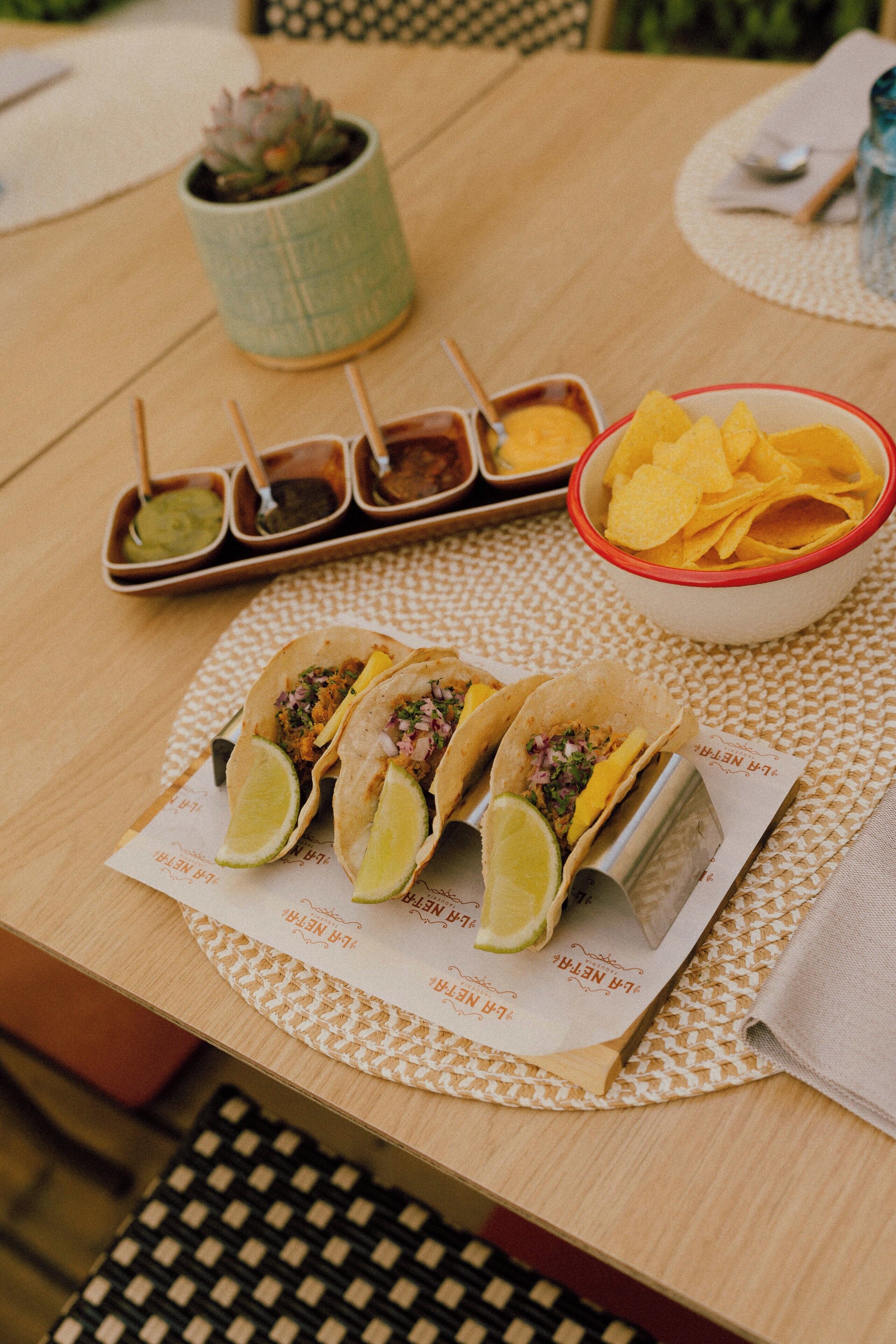 A table with three tacos, a bowl of chips, and four small containers of sauces.