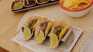 A table with three tacos, a bowl of chips, and four small containers of sauces.