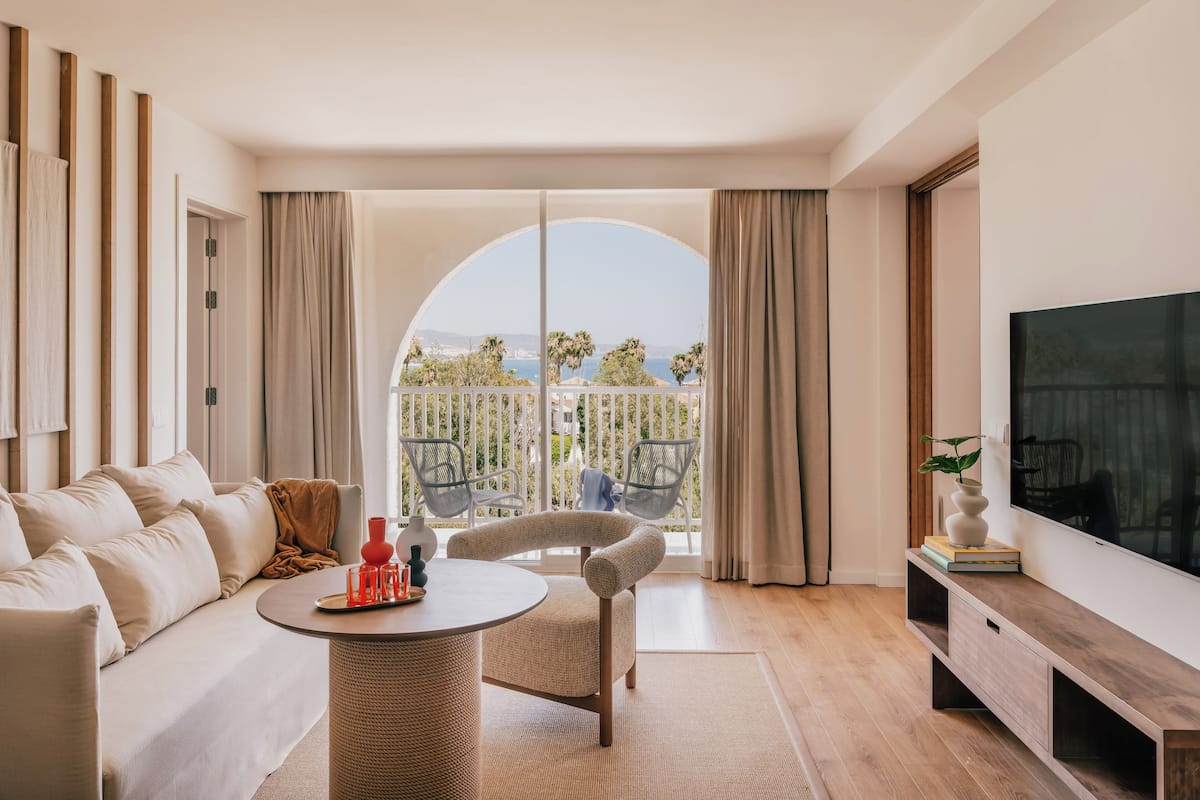 A modern living room with a sofa, a round table, and a TV. There is a balcony with a view of the sea and trees.