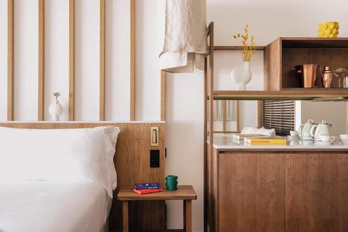 A hotel room with a bed, a bedside table with books and a green mug, a wooden nightstand, a mirror, and a shelf with kitchen items.