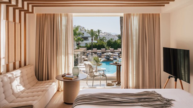 Une chambre d’hôtel avec un lit, un canapé, une petite table et un téléviseur. La fenêtre donne sur une piscine et un bâtiment.