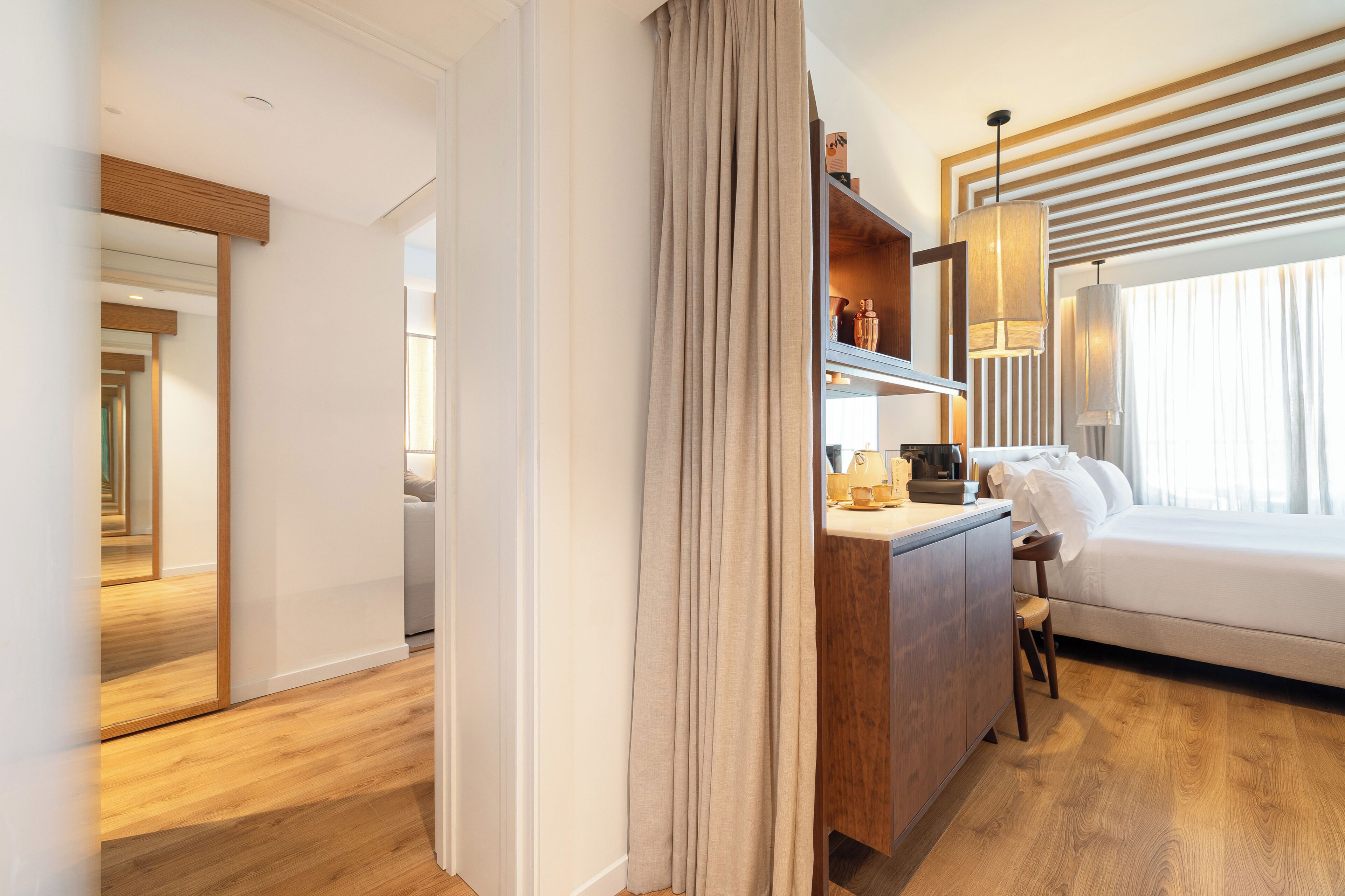 A hotel room with a wooden floor, a bed with white bedding, a desk with a lamp, and a large window with curtains.