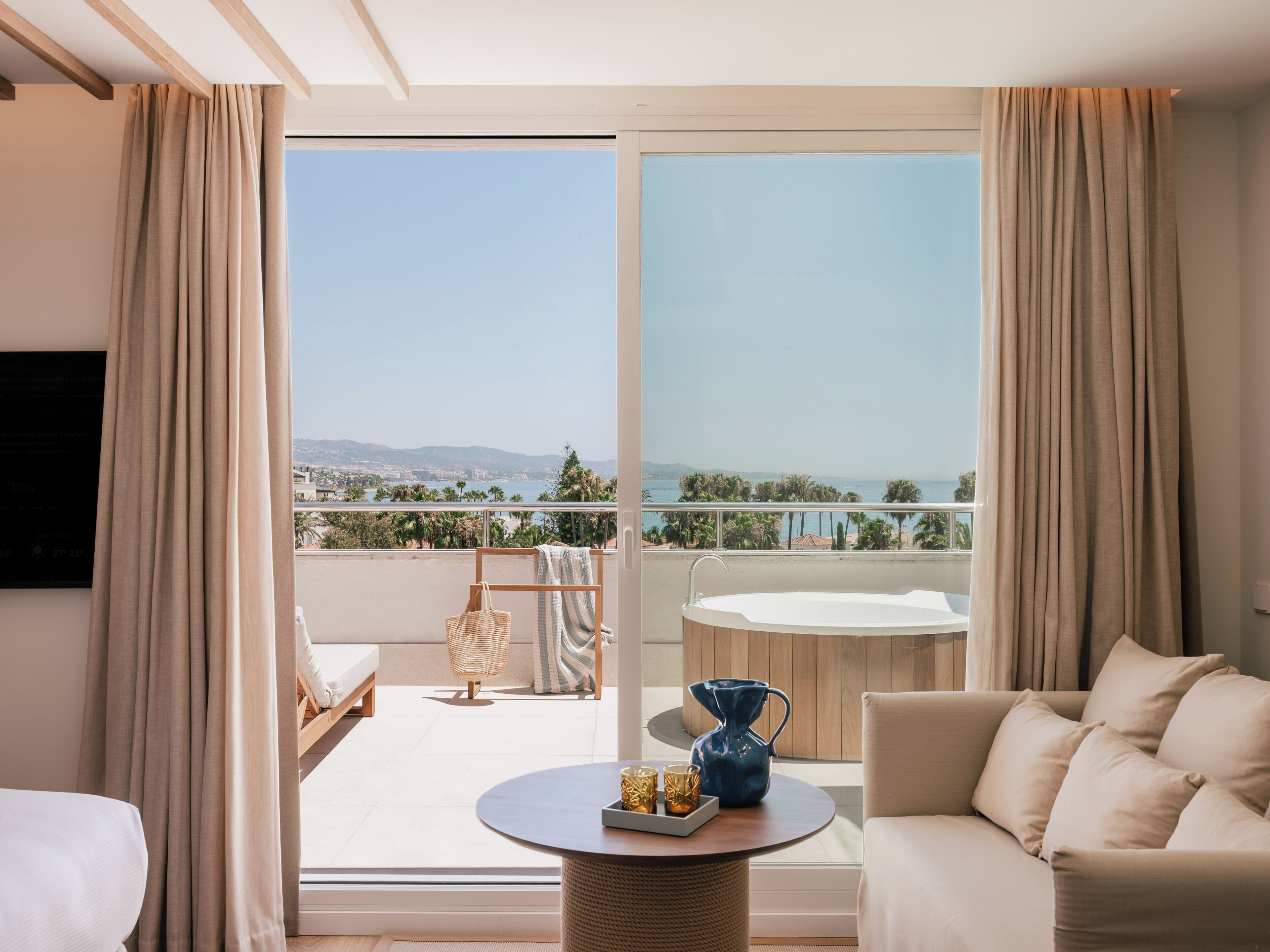 Une chambre d’hôtel moderne dotée d’un balcon, d’un canapé, d’une table basse et offrant une vue sur la mer et les collines.